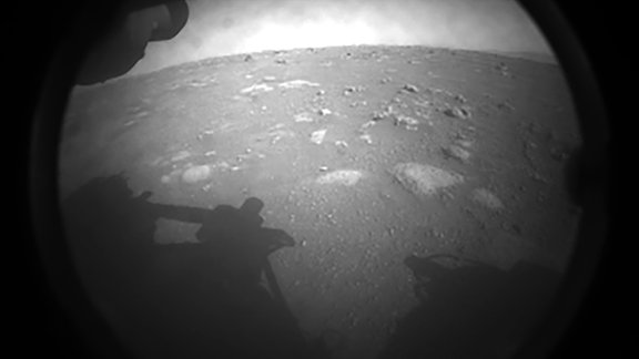 Eine schwarz-weiß Aufnahme der Marsoberfläche. Man erkennt den Schatten des Rovers und die staubige Oberfläche des Mars. An einigen stellen befinden sich flache Steine.