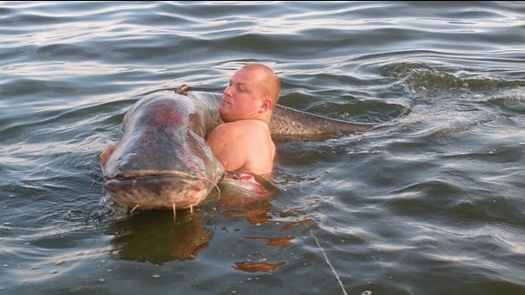 Ein Mann steht im Wasser und hält einen großen Fisch in den Armen.