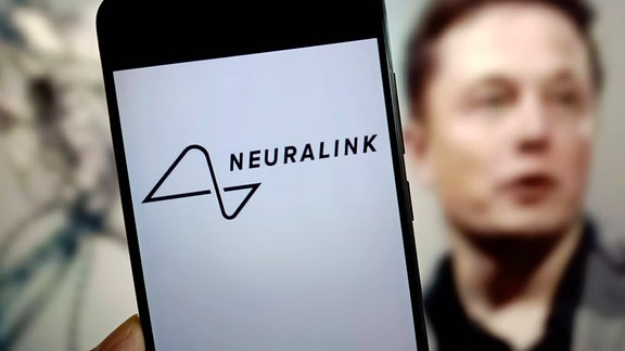 Handy mit Logo von Neuralink auf Bildschirm, im Hintergrund unscharf Porträt von Elon Musk, der zur Seite blickt