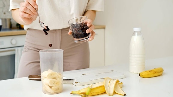 Frau gibt schwarze Johannisbeeren in einen Mixer und mischt sie mit einer zerkleinerten Banane, im Hintergrund eine Flasche Milch