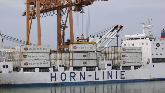 Containerschiff im Hafen mit Aufschrift "Horn-Linie" und gestapelten Container für Obsttransport. Containeraufschrift Logo und englischer Text "Say yes to the best". Großer Kran verlädt Container.