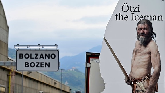 Werbedarstellung in Bozen zeigt Ötzi als älteren Steinzeit-Mann mit hellerer Hautfarbe und längerem Haar. Links vermutlich Bahnhofschild "Bolzano, Bozen", im Hintergrund Berge.
