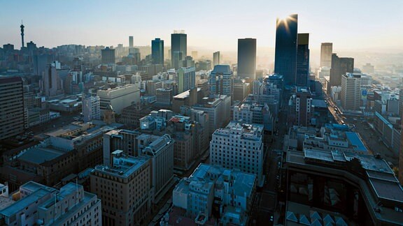 Skyline von Johannesburg mit dichter Innenstadtbebauung, Hochhäusern bzw. Wolkenkratzern, tief stehender Sonne als Blendflecken nahe am höchsten Hochhaus, blauer Himmel mit gelblichem Streifen Richtung Horizont.