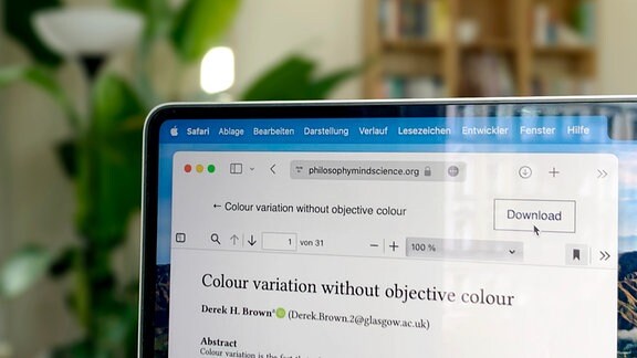 Laptopbildschirm mit offenem Browserfenster zeigt einen Artikel im Journal Philosophy and the Mind Sciences mit Mauszeiger Download-Button. Im Hintergrund unscharf Regal und große grüne Zimmerpflanze.