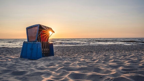 Einzelner Strandkorb im Sand, darin Gegenlicht von untergehender Sonne, Meer im Hintergrund.