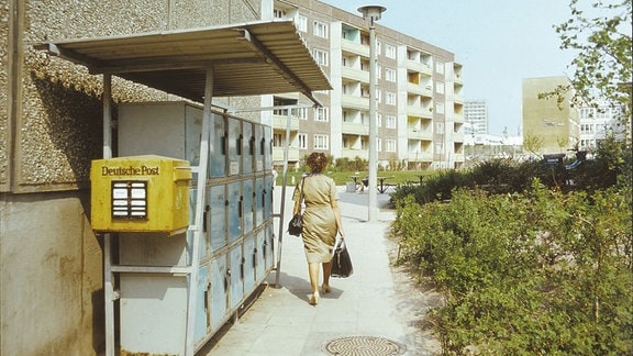 Eine Frau geht an einer HAuswand entlang, davor hängt ein gelber Postkasten und dahinter gibt es eine Reihe von Schließfächern