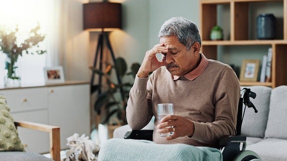 Ein Senior in einem Rollstuhl stützt müde seinen Kopf und hält ein Wasserglas.