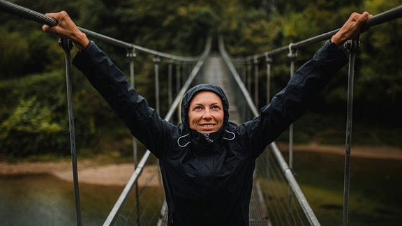 junge Frau zentral im Bild auf einer Hängebrücke mit Regenjacke und aufgezogener Kapuze bei Regenwetter, hält sich links und rechts an Geländer fest und blickt nach oben