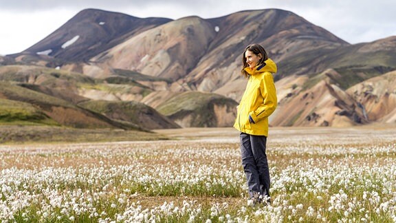 junge Frau mit gelber Regenjacke steht auf Blumenwiese, im Hintergrund sanft kurvige karge Berge in Island