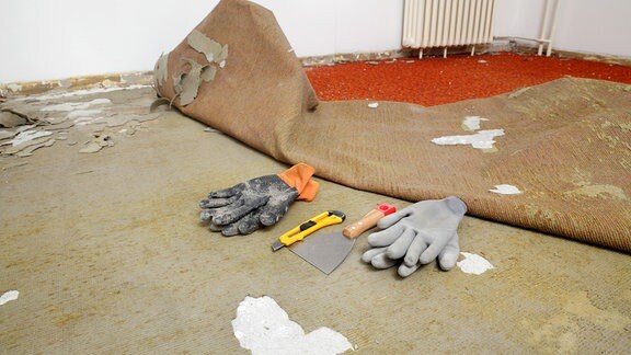 in einem Zimmer liegen Handschuhe, Spachtel und ein Messer neben einem roten Teppichboden, der bereits teilweise herausgerissen ist, im Hintergrund Heizkörper