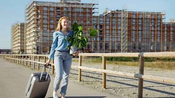 Junge Frau geht mit Pflanze und Rollgepäck an einem sonnigen Tag spazieren