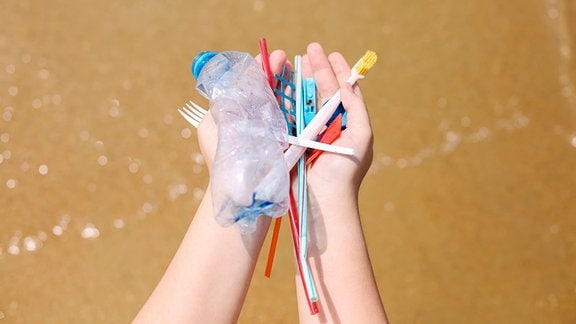 Hand hält über Wasser verschiedene gefundene Plastikgegenstände wie Wasserflasche, Zahnbürste, Gabel, Strohhälme.