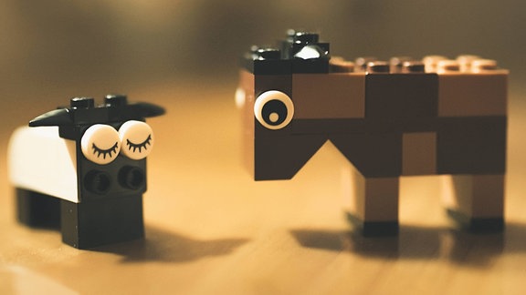 Nahaufnahme von zwei Tieren aus Klemmbausteinen wie Lego, eventuell Schaf und Esel, in gedeckten Farben (braun, weiß), Stehen auf Tisch, Hintergrund unscharf. Schaf mit geschlossenen, Esel mit offenen Augen.