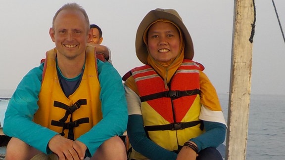 Ein Mann und eine Frau mittleren Alters sitzen nebeneinander auf einem Holzboot. Sie tragen Schwimmwesten. 