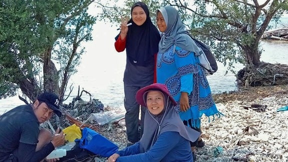 Drei Frauen und ein Mann stehen auf einem Strand mit Plastikmüll. 