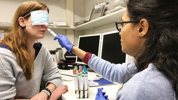 Podcastreporterin Daniel Schmidt hat die Augen verbunden während ihr eine Forscherin eine Geruchsprobe unter die Nase hält.