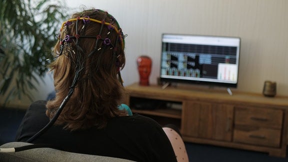 Eine Frau sitzt in einem Raum und schaut auf einen Fernsehbildschirm. Ihr Kopf ist mit einer Sensorenhaube verkabelt. Dabei handelt es sich um eine Neurofeedback-Therapiesitzung