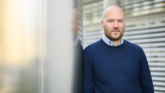 Das Foto zeigt einen Mann in Hemd und Pullover, der an eine Wand gelehnt in die Kamera blickt.