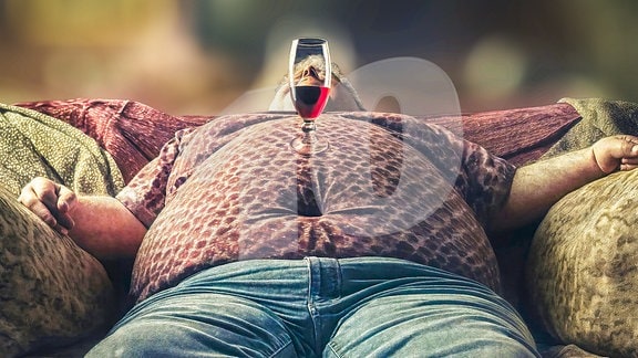 Korpulenter Mann liegt auf einem Sofa und schläft, auf dem Bauch ein Weinglas balancierend.