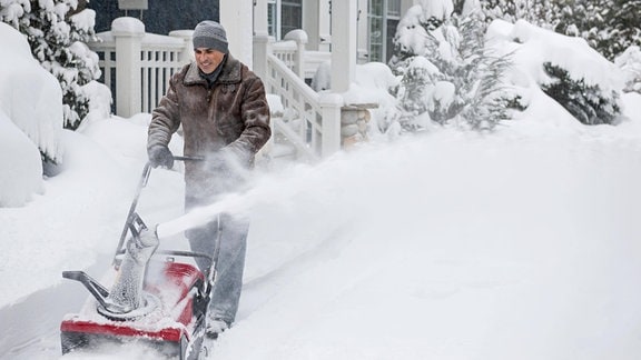 Ein Mann räumt nach starkem Schneefall mit einer Schneefräse den tiefen Schnee auf der Einfahrt zu einem Wohnhaus.