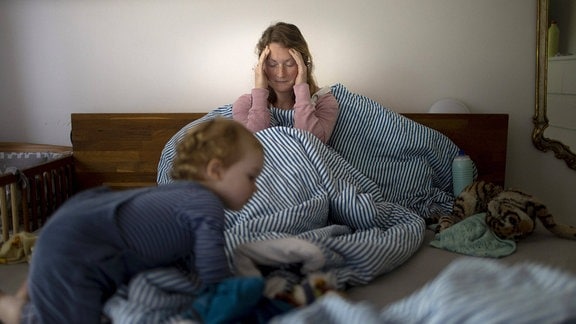 Eine Mutter hält sich den Kopf, während ein Kleinkind über ihr Bett krabbelt.