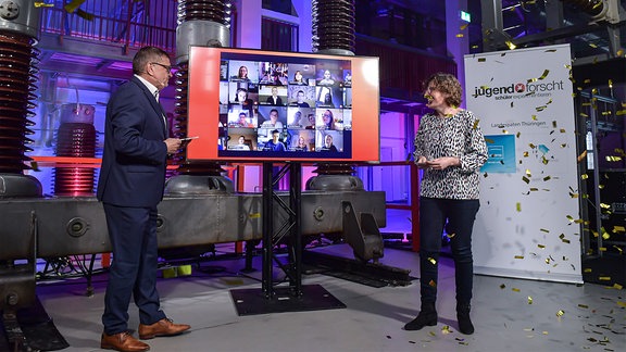 Ein mann und eine Frau moderieren eine Veranstaltung – in der Mitte ein Bildschirm mit vielen Webcambildern aus Videoschalten
