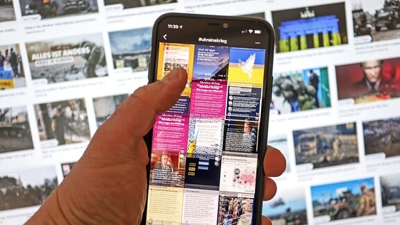 News bei Instagram auf einem Smartphone in einer Hand werden durchgescrollt. News zum Krieg in der Ukraine. Im Hintergrund Nachrichtenbilder, Bilder auf einem PC-Monitor zum Krieg in der Ukraine.