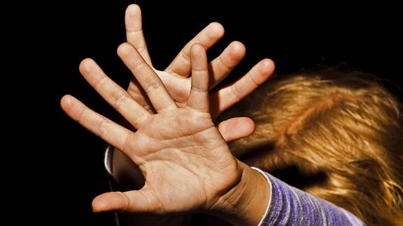 Symbolbold: Mädchen streckt Hände abwehrend in richtung Kamera, Kopf gesenkt und kaum zu erkennen, Hintergrund dunkel