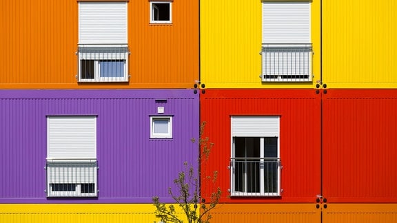 Container in grellem Orange, Gelb, Lila und Rot sind aufeinandergestapelt und ergeben zusammen eine bunte Fassade. In Jedem Container gibt es ein großes Fenster. In drei von vier Fenstern ist der Rollladen runtergelassen. 