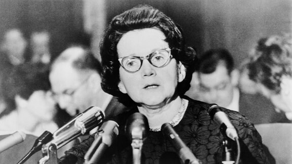 historisches Bild: Eine Frau mit Brille an einem Rednerpult