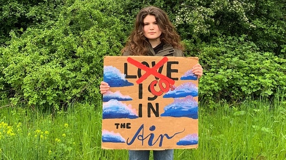 Eine junge Frau mit gelockten braunen Haaren. Vor ihrem Körper ein großes Schild mit der Aufschrift "Love is in the air", auf der das Wort "Love" durchgestrichen und durch CO2 ersetzt wurde.