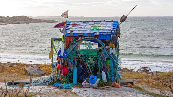 Eine kleine Hütte, die aus verschiedenen bunten Plastikmüllteilen (z.B. Flaschen und ein Netz) zusammengebaut wurde, an einem Strand. Im Hintergrund Wasser bis zum Horizont.