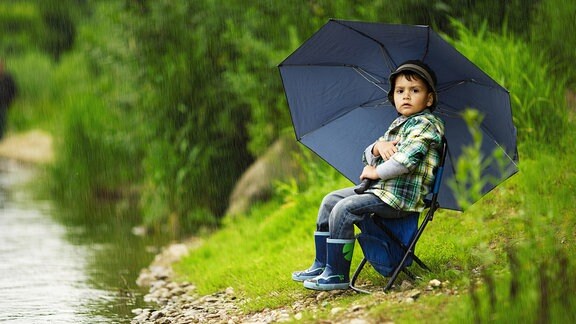 Kleiner Junge  mit Regenschirm sitzt auf einem Klappstuhl