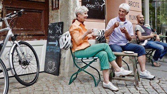Ältere Menschen trinken Kaffee vor einem Szenecafé, neben ihnen Elektrobikes.