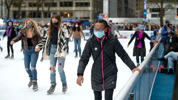 Mädchen mit unterschiedlicher Hautfarbe und Mund-Nasen-Bedeckung auf einer Eisbahn beim Schlittschuhlaufen in Manhattan. Im Hintergrud unscharf viele Menschen und Gebäude.