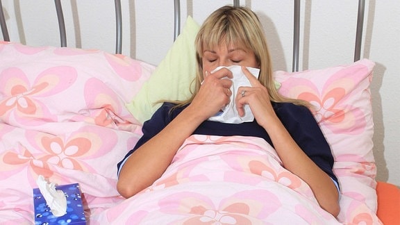 Frau liegt erkältet im Bett und putzt sich die Nase.