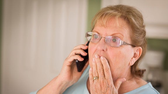 Eine ältere Frau telefoniert und fasst sich erschrocken an den Mund.