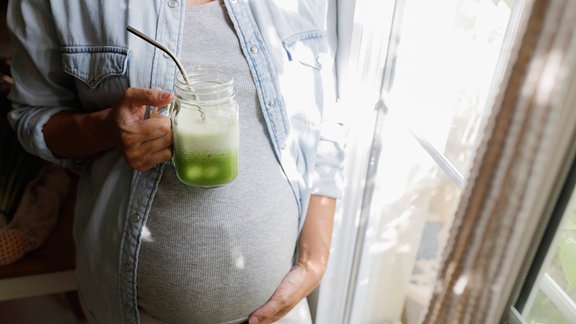 Eine Schwangere Frau mit grünem Smoothie-Glas am Fenster zu Hause.