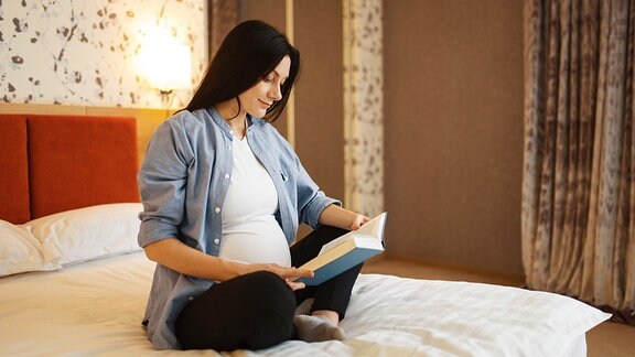 Eine schwangere Frau sitzt mit einem Buch auf einem Bett