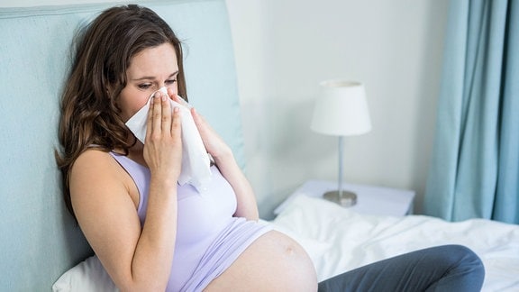 Eine schwangere Frau sitzt im Bett und putzt sich die Nase