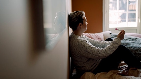 Distanziert aufgenommenes Bild: Frau mit kurzen Haaren sitzt in Schlafzimmer auf Bett an Wand gelehnt, hält Schwangerschaftstest in der Hand und schaut aus Fenster. Warme, entsättigte Farben, Vorder- und Hintergrund unscharf.