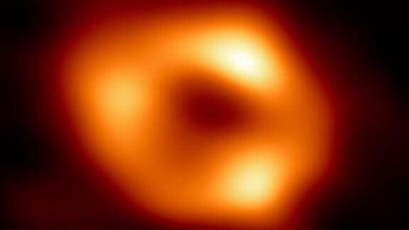 Das erste Bild des schwarzen Lochs Sagittarius A*: Ein orangener Ring mit hellen gelben Stellen und dem schwarzen Schatten des Lochs in der Mitte vor dunklem Himmel.