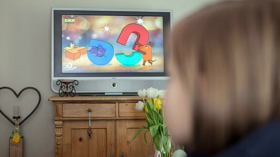 Ein Kind sitzt im Wohnzimmer auf einer Couch und schaut die Sendung mit der Maus.