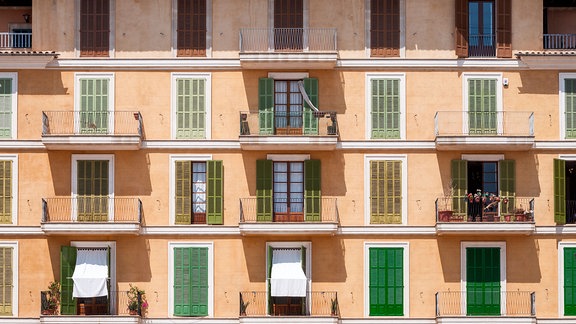 Die Fassade eines Wohnhauses mit Balkonen und Balkontüren, die in der Mittagssonne teilweise mit Rollläden geschlossen sind.