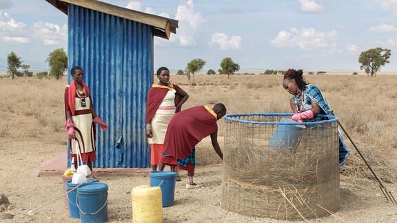 Vier Frauen arbeiten mit Eimern in einer steppenartigen Umgebung vor einem blauen Toiletten-Häuschen
