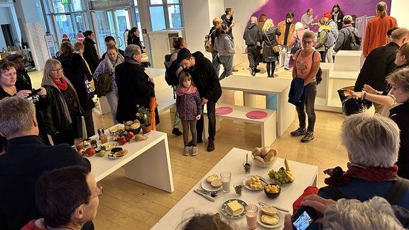 Menschen besuchen eine Ausstellung, auf Tischen stehen Zutaten für ein Frühstück.