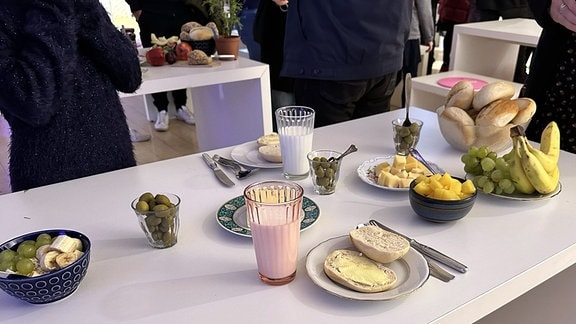 Auf einem Tisch stehen Gläser mit Oliven, zwei aufgeschnittene und mit Butter geschmierte Brötchen, sowie zwei Gläser mit weißer Flüssigkeit.