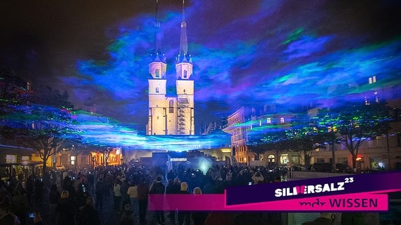 Besucher bei Nacht auf dem Marktplatz in Halle, im Hintergrund die Marktkirche, darüber eine Lichtinstallation.