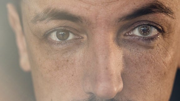 Frontale Nahaufnahme eines dunkelhaariger Mannes mit hochgezogener Augenbraue und Kontaktlinse, die wie eine digitale Kontaktlinse aussieht. Mystischer Lichteinfach und Unschärfe von links.