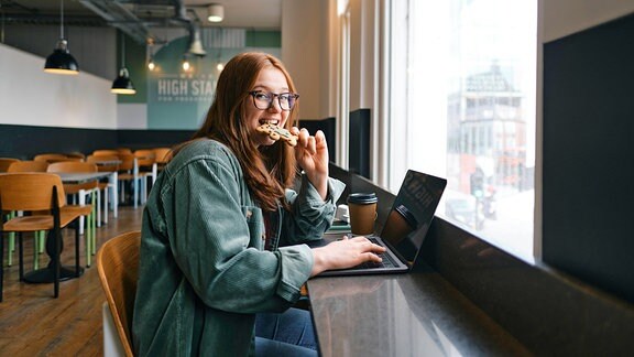 Eine junge Frau sitzt am Fenster eines Cafés vor einem Laptop und isst etwas